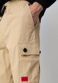 Beige cargo shorts laget av lett stoff, med sidelommer med svarte knapper og en rød logo-patch på nedre kant.