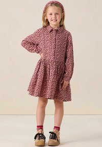 Robe rose à imprimé léopard avec des manches longues, un col et une jupe froncée. Associée à un serre-tête assorti et des chaussures beiges avec des lacets noirs.