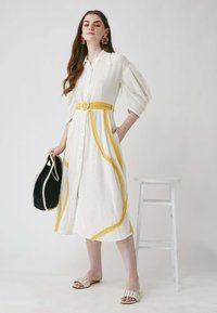 Weißes Hemdkleid mit gelben Akzenten, übergroßen Ärmeln und einem taillierten Gürtel. Der Stoff hat eine strukturierte Oberfläche. Getragen mit einer schwarzen Tasche und Sandalen.