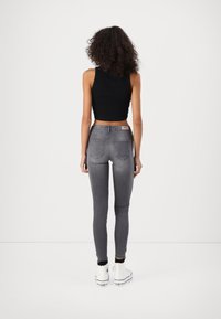 Mujer con cabello rizado, vistiendo una camiseta corta negra, jeans ajustados grises y zapatillas altas blancas, de pie con la espalda hacia la cámara.