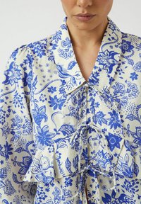 Vit blus i tyg med intrikata blå blommönster och paisleymönster, med en volangslängd, krage och knytning fram.