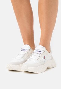 Białe tenisówki z mieszanką gładkiej i zamszowej tekstury. Charakteryzują się grubymi podeszwami, zaokrąglonymi noskami oraz logo na języku i boku.