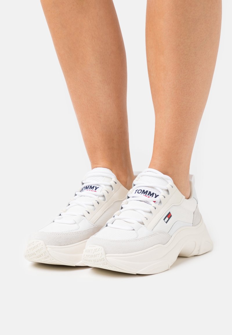 Białe tenisówki z mieszanką gładkiej i zamszowej tekstury. Charakteryzują się grubymi podeszwami, zaokrąglonymi noskami oraz logo na języku i boku.