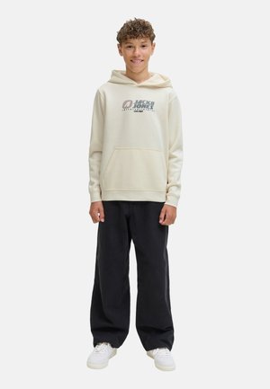 Ein lächelnder junger Mensch steht in einem beigen Hoodie mit Logo, kombiniert mit schwarzen Hosen und weißen Turnschuhen, vor einem schlichten Hintergrund.