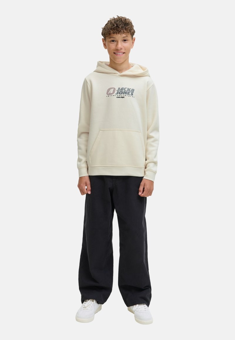 Une jeune personne souriante se tient dans un hoodie beige avec un logo, associé à un pantalon noir et des baskets blanches, devant un fond uni.