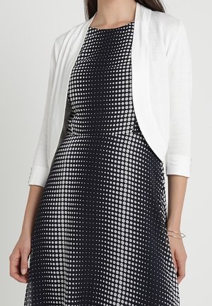 Femme portant une robe noire à motif de pois blancs et un cardigan court blanc à manches trois-quarts sur un fond uni.