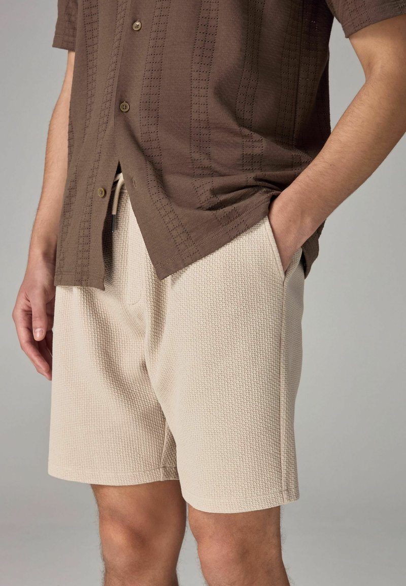 Mand iført beige strukturerede shorts og en brun kortærmet skjorte med knapper, den ene hånd i lommen, foran en ensfarvet grå baggrund.