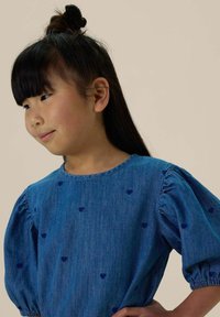 Robe en denim bleu clair avec des manches bouffantes, présentant de petits motifs de cœurs marins sur l'ensemble du tissu. Encolure ronde et texture douce.