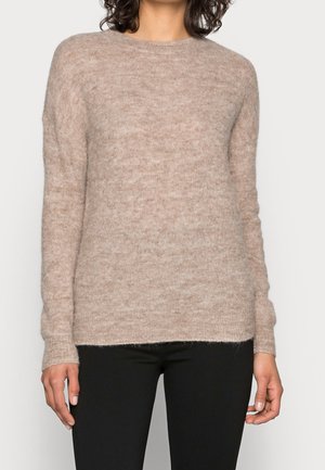 Kvinde iført en beige langærmet, fluffy sweater kombineret med sorte bukser, stående mod en ensfarvet lys baggrund.