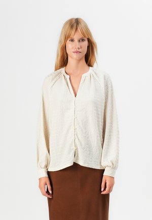 Blonde vrouw in een textuur off-white blouse met knopen en lange mouwen, met een bruine rok, staand tegen een effen witte achtergrond.