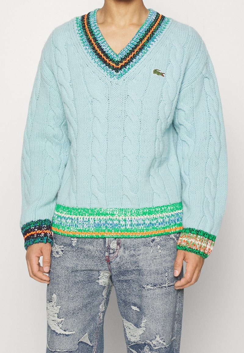 Ljusblå kabelstickad tröja med V-ringning, som har flerfärgade randiga detaljer vid halsringningen och ärmarna. Ribbad nederkant.