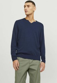 Maglione lavorato a maglia blu navy con un sottile scollo a V, maniche lunghe e polsini a coste. Abbinato a pantaloni chino verde chiaro.