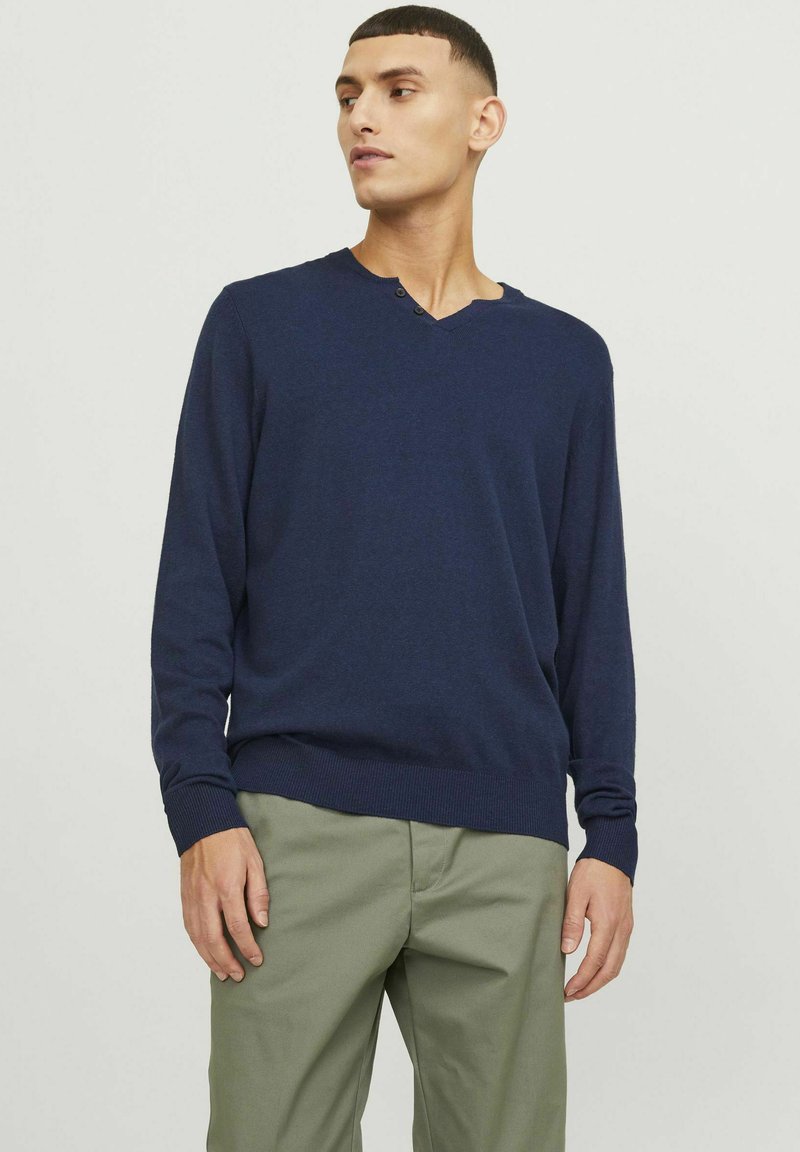 Maglione lavorato a maglia blu navy con un sottile scollo a V, maniche lunghe e polsini a coste. Abbinato a pantaloni chino verde chiaro.