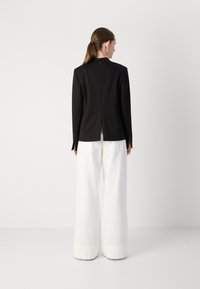 Blazer nero con maniche lunghe e uno spacco centrale sul retro, abbinato a pantaloni bianchi a gamba larga. Tessuto liscio, design minimalista, senza chiusure visibili.