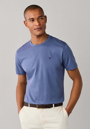 Man draagt een blauw t-shirt met ronde hals en een klein logo op de borst, beige broek en een bruine geweven riem, handen in de zakken, kijkt naar links.