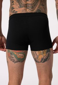 Boxer shorts en coton noir à coupe ajustée, avec une ceinture douce et des tatouages visibles sur les cuisses.
