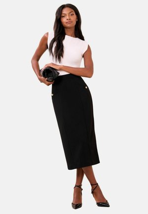 Woman wearing sleeveless white top and black midi skirt with gold buttons, holding black clutch, wearing black pointed heels, standing.