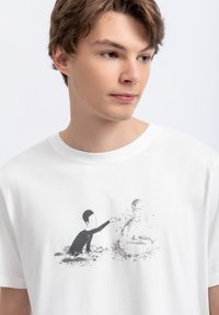 Witte katoenen t-shirt met een zwart cartoonafbeelding van een kind dat reikt naar een figuur gemaakt van verspreide deeltjes. Korte mouwen.