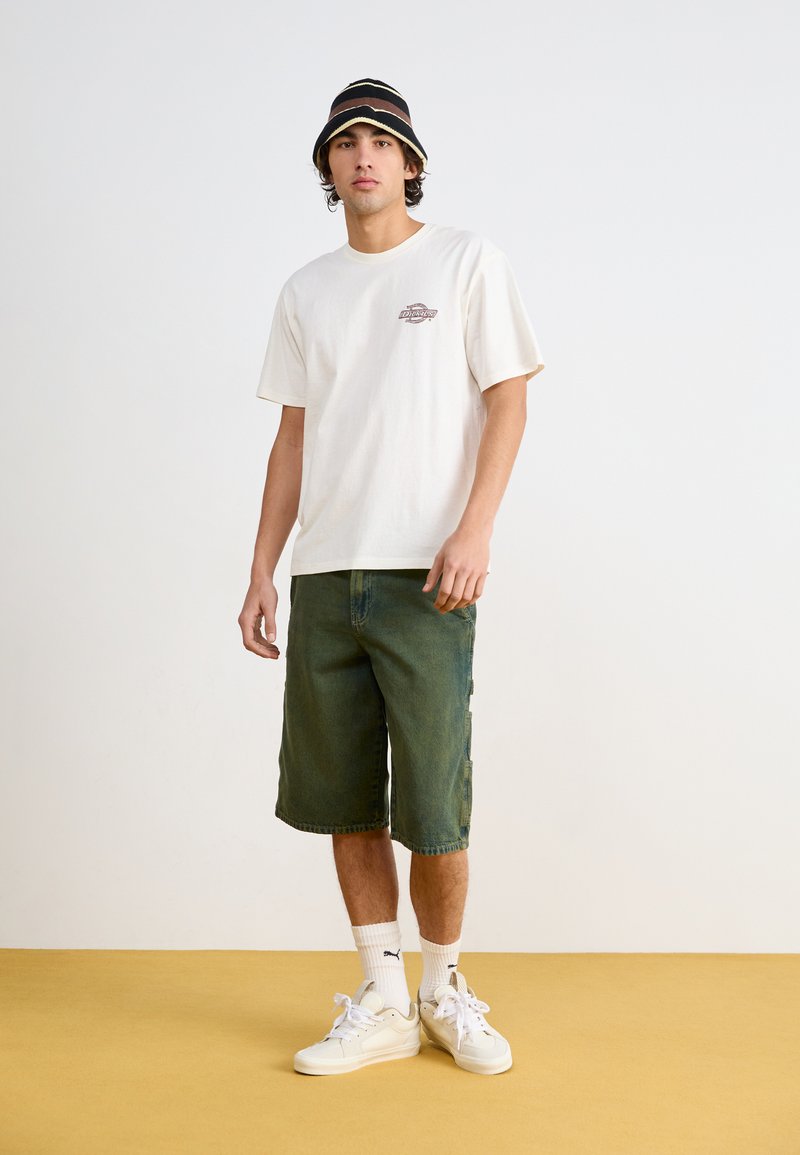 T-shirt bianca in cotone con un piccolo logo, pantaloni cargo in denim verde, sneakers bianche e un cappello a secchiello a righe nere e marroni, su un pavimento giallo.