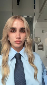 Camicia azzurro chiaro con colletto appuntito, cravatta scura con motivo, e lunghi capelli biondi ondulati. Sfondo neutro con elementi di bagno.