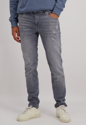 Slim-fit grijze denim jeans met versleten details en vervaagde gebieden. Combinatie met witte sneakers en een blauwe sweatshirt.