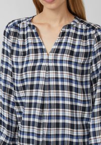 Kariertes Hemd mit einem navyblauen, blauen und weißen Karomuster sowie roten Akzenten. V-Ausschnitt und geraffte Details an den Schultern.