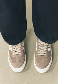 Brune ruskind sportssko med hvide accenter og snørebånd. Runde tå og tekstureret sål. Båret med sorte bukser mod en neutral baggrund.