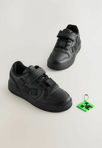 Zwarte sneakers met een gladde leren afwerking, voorzien van klittenbandsluitingen, geperforeerde neus en een pixelachtige groene en zwarte sleutelhanger eraan bevestigd.