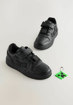Zwarte sneakers met een gladde leren afwerking, voorzien van klittenbandsluitingen, geperforeerde neus en een pixelachtige groene en zwarte sleutelhanger eraan bevestigd.