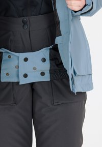 Blå isolerad jacka med dragkedja, svarta knappar och hög krage, i kombination med svarta snöbyxor med stora fickor och en mjuk textur.