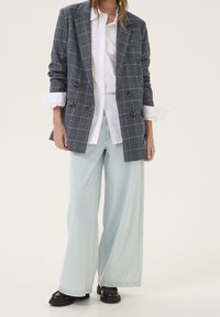 Marineblauer doppelseitiger Blazer mit Karomuster über einem weißen Hemd, kombiniert mit hellblauen Jeans mit weitem Bein und schwarzen Schuhen. Glatte Texturen und maßgeschneiderte Passform.