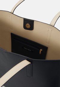 Black tote bag with beige interior, featuring a zippered pocket, gold hardware, and two contrasting handles. Smooth texture.