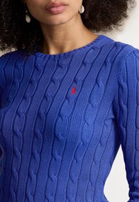Blå stickad tröja med ett kabelstickat mönster och en liten röd logotyp på bröstet. Rund halsringning med en strukturerad yta.