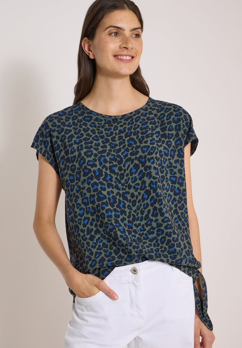 Femme souriante portant un haut à manches courtes bleu et noir à imprimé léopard avec un nœud sur le côté et un pantalon blanc, main dans la poche, sur fond uni.