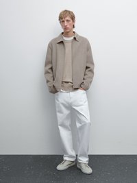 Beige uldjakke med en afslappet pasform, parret med en lysebeige sweater og hvide bukser, suppleret med lysfarvede sneakers.
