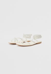 Sandales plates blanches avec plusieurs brides croisées, ornements scintillants et une fine sangle de cheville, posées sur une semelle marron clair.