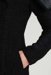 Černá pletená bunda s texturovaným vzorem, bočními kapsami a volným střihem s mírně oversized rukávy.