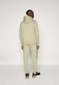 Homme avec des cheveux tressés portant un sweatshirt à capuche vert clair et un pantalon de jogging assorti avec des bandes latérales, des baskets Puma noires et vertes, se tenant de dos.