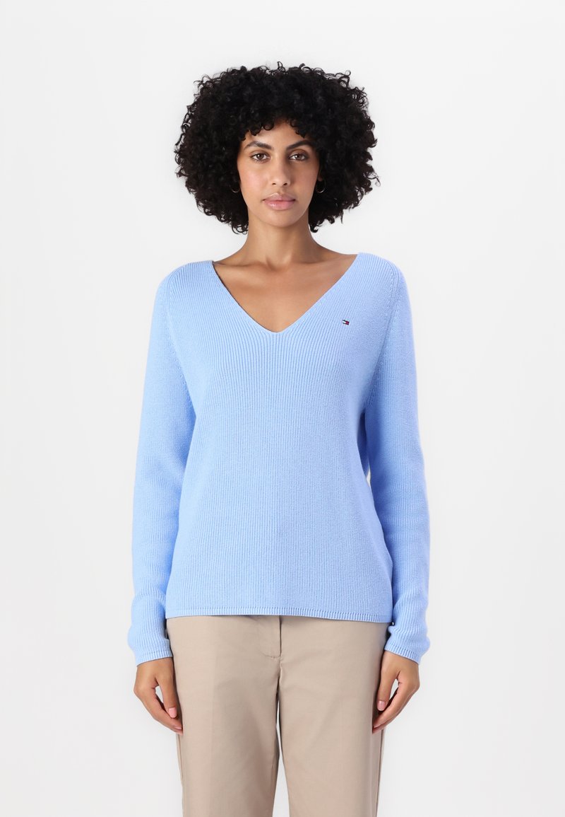 Svetlo modra v-neck pulover s rebrasto teksturo, dolgi rokavi in majhna majica na levi rami, v kombinaciji z bež hlačami.