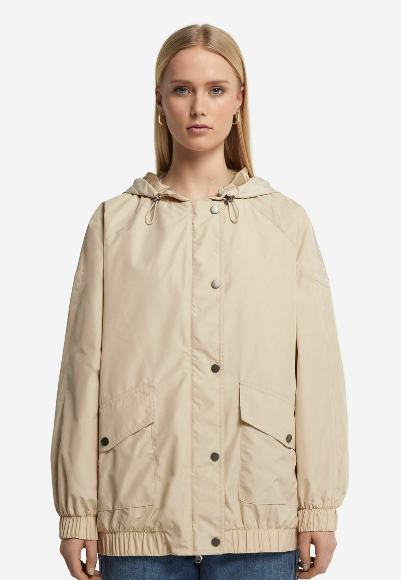 Beige huvmantel med en lätt design, tryckknappar, elastiska ärmslut och två framfickor. Slät textur och avslappnad passform.