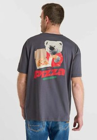Man met een donkergrijze T-shirt met een afbeelding van een beer in een rode jas die een pizzastuk vasthoudt en het woord "Pizza" in rode tekst.