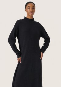 Rochie lungă neagră cu mâneci lungi și guler înalt, realizată dintr-un material texturat moale. Prevăzută cu slituri laterale pentru ușurința mișcării.