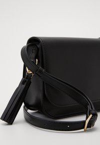 Černá kožená kabelka přes tělo s hladkým povrchem, zlatými kovovými akcenty a detaily s třásněmi na popruhu. Obsahuje překrytí s klapkou.