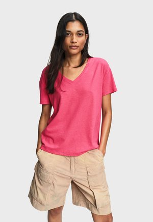 Femme aux longs cheveux bruns portant un t-shirt rose à col en V et un short cargo beige, debout les mains dans les poches devant un fond uni.