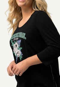 Camiseta de algodón negra con escote redondeado, adornada con bordados florales y el texto "Sociedad Horticultural". Mangas tres cuartos y textura suave.