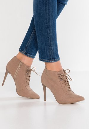 Bottes cheville à talons hauts en daim marron clair avec lacets et fermeture éclair latérale, portées avec un jean skinny bleu sur fond blanc.