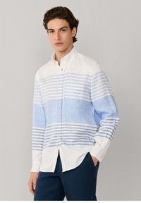 Joven con cabello oscuro que lleva una camisa de manga larga abotonada con rayas horizontales blancas y azul claro, pantalones oscuros, con la mano en el bolsillo.