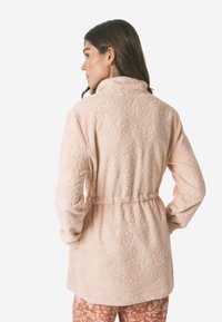 Lyserød fleece morgenværd med blomsterdesign, krave og bælte i taljen. Blødt materiale, lange ærmer og midtlange design, der går til låret.