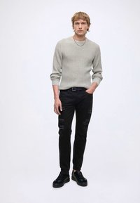 Uomo in piedi che indossa un maglione a coste grigio chiaro, jeans skinny neri strappati, cintura nera, sneakers nere, con una mano in tasca.