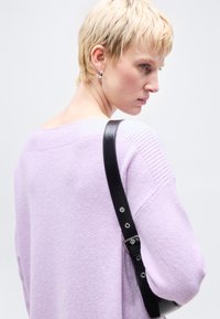 Γυναίκα με κοντά ξανθά μαλλιά, που φοράει ασημένια σκουλαρίκια, λιλά πουλόβερ και μαύρο λουράκι τσάντας στον ώμο, μπροστά από ένα απλό φωτεινό φόντο.